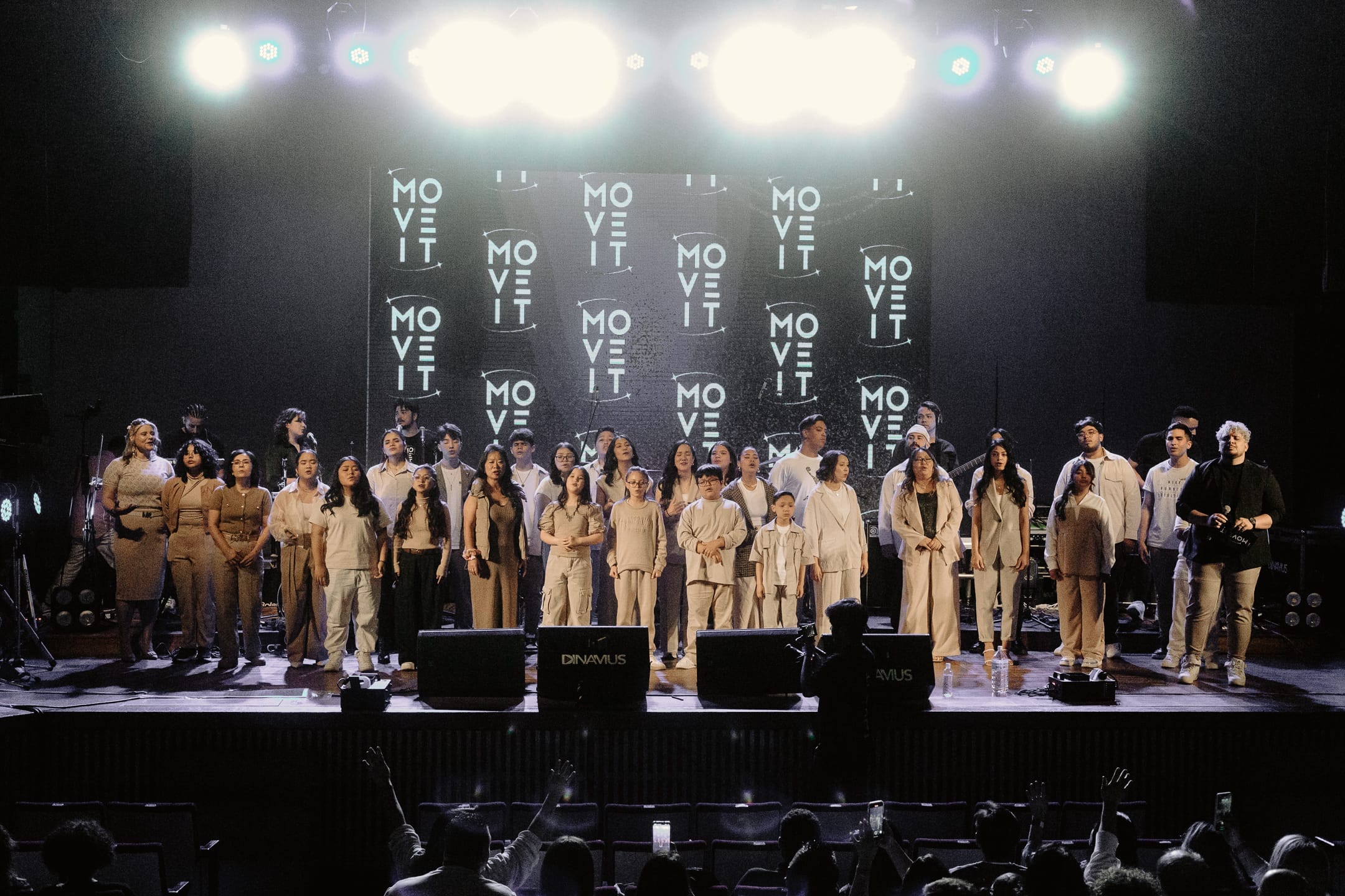 Palco cheio do recital Dias Music, coral e banda se apresentando sob luzes de show